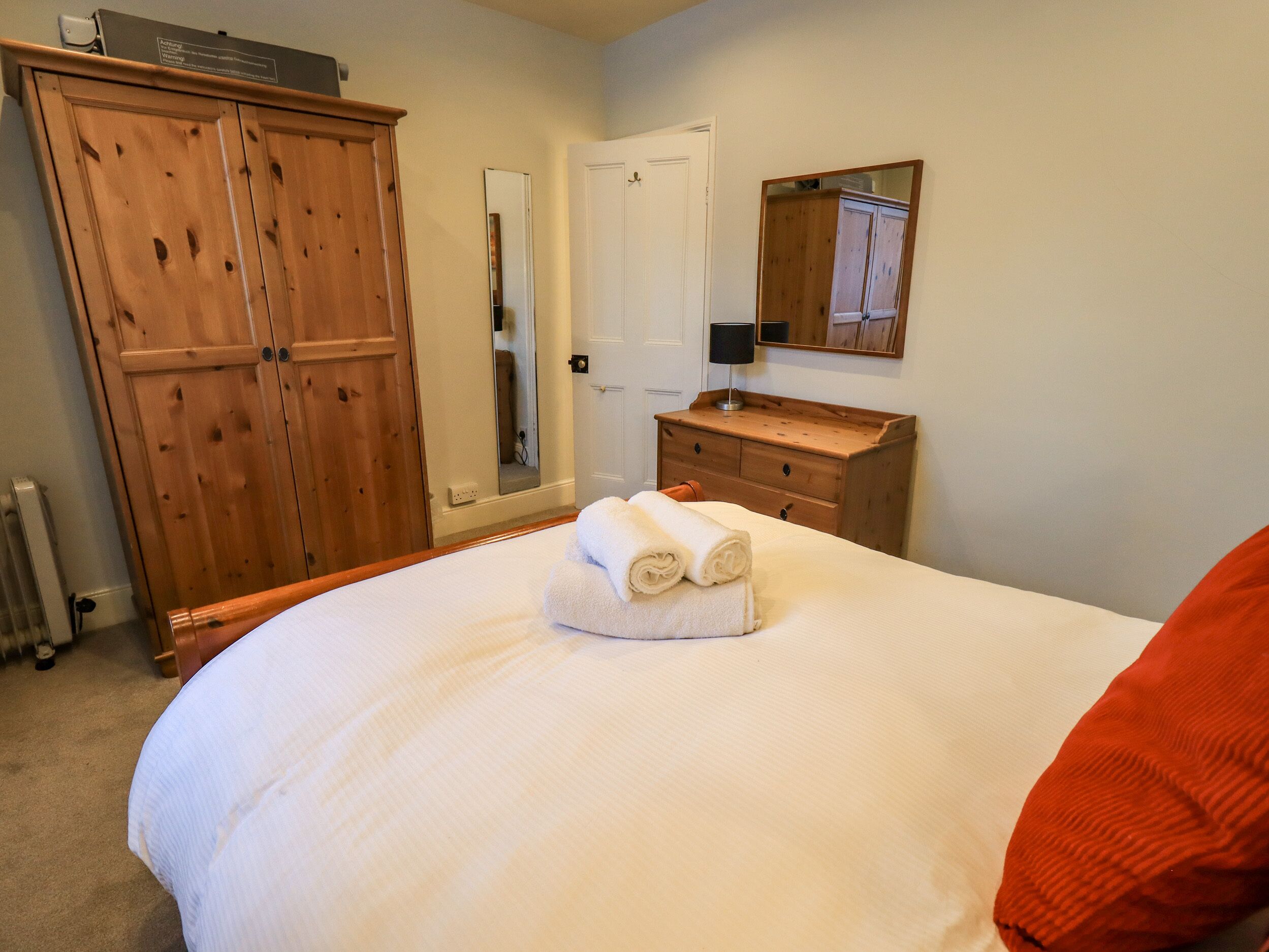 A bedroom with a large bed, wooden wardrobe, a mirror, and a small dresser.