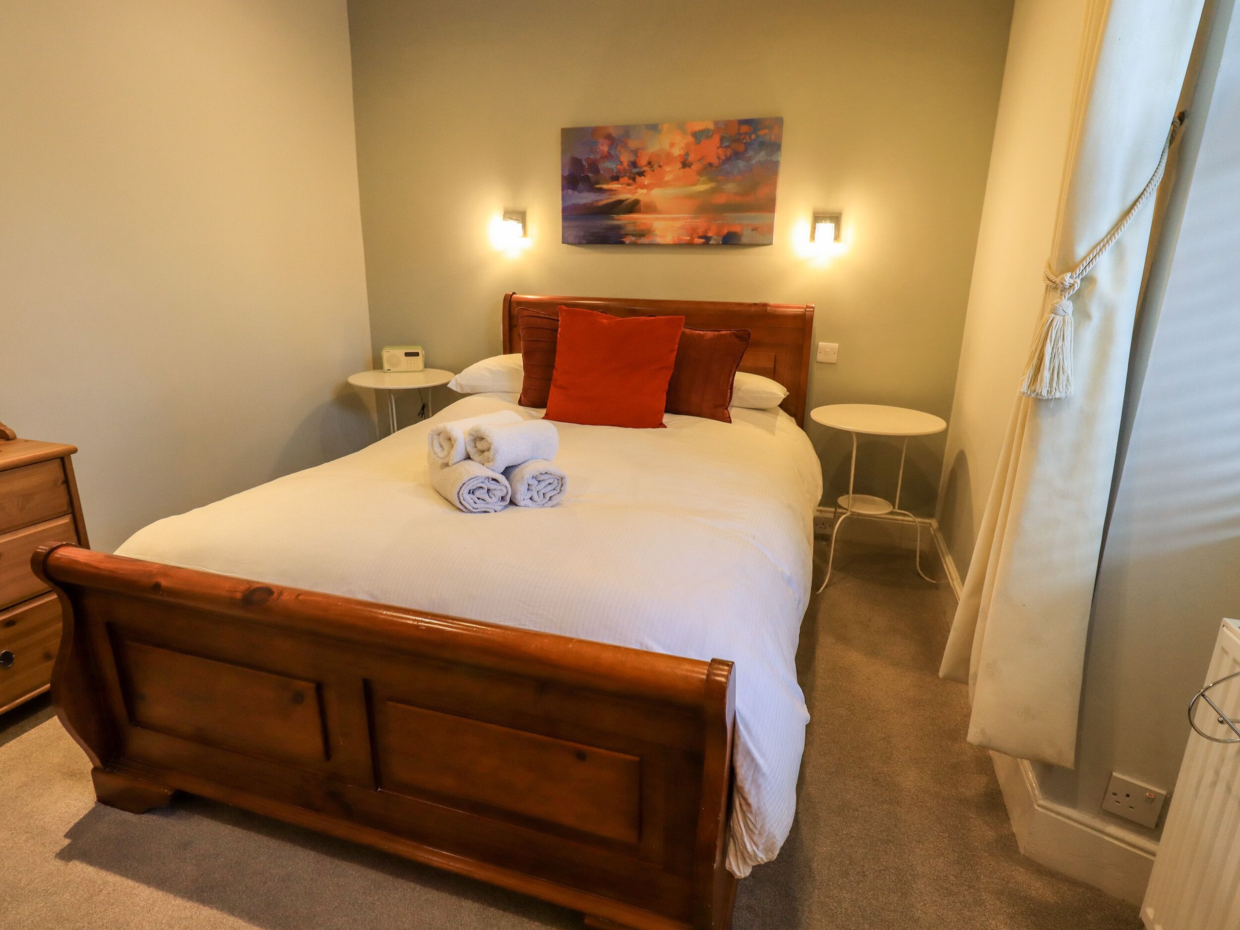 A neatly made bed with a wooden headboard, a painting on the wall, and a small bedside table with a lamp.