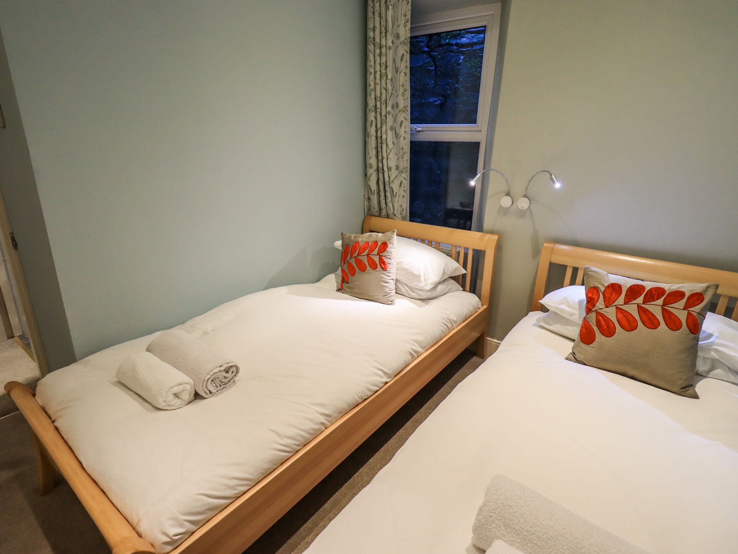 A twin bedroom with a wooden bed frame, two pillows with leaf patterns, a bedside lamp, and a window with a view of trees.