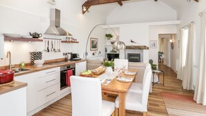 A spacious kitchen with wooden beams, a dining area with white chairs, and a cozy fireplace. - Beudy Hywel (Caernarfon)