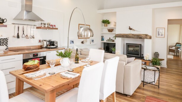 A modern kitchen with wooden beams, a dining area with a wooden table, white chairs, and a fireplace. - Beudy Hywel (Caernarfon)