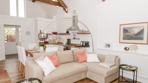 A modern living room with a beige sectional sofa, a small table, and a kitchen area with a hanging light fixture. - Beudy Hywel (Caernarfon)