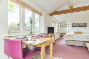Quarto com mesa de jantar, cadeiras, televisão, cama e janela com vista para uma parede de pedra e vegetação.