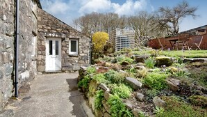 Um edifício de pedra com uma porta branca, um jardim com pedras e plantas, e uma cerca de madeira.