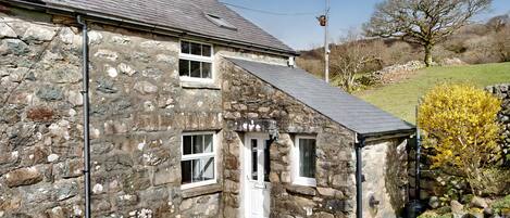 A stone house with a sloped roof, a small porch, and a garden with flowers and a tree.