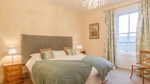Um quarto com uma cama grande, uma mesa de cabeceira de madeira, uma cadeira, um candelabro e uma janela com cortinas.