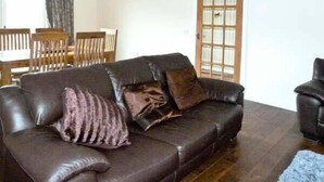 A living room with a dark brown leather sofa, a wooden dining table with chairs, and a wooden door with glass panels. - Braewood (Inverness)