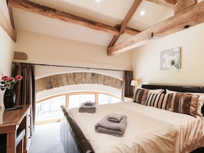 A bedroom with a large bed, wooden beams, a stone arch window, and a vase with flowers on a bedside table. - Stoneycroft Barn (Sheffield)