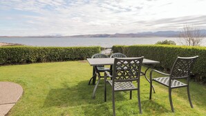 Un área de estar al aire libre con mesa y sillas, un seto y vistas a un lago y montañas.