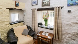 A cozy living room with a sofa, a small table with a vase of flowers, and a window with curtains. There are framed pictures on the wall and a lamp on a side table. - Ffynnon Tom (Haverfordwest)