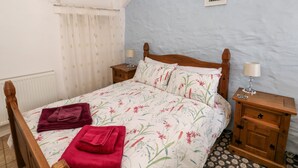 A bedroom with a wooden bed, floral bedding, two bedside tables with lamps, a radiator, and a window with curtains. - Ffynnon Tom (Haverfordwest)