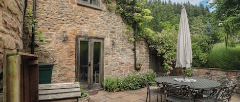 A stone building with a wooden bench, a table with chairs, and a garden with flowers.