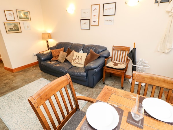 A living room with a blue leather sofa, wooden chairs, a dining table with plates and a glass, and wall-mounted artwork. - Hunter Cottage (Haltwhistle)