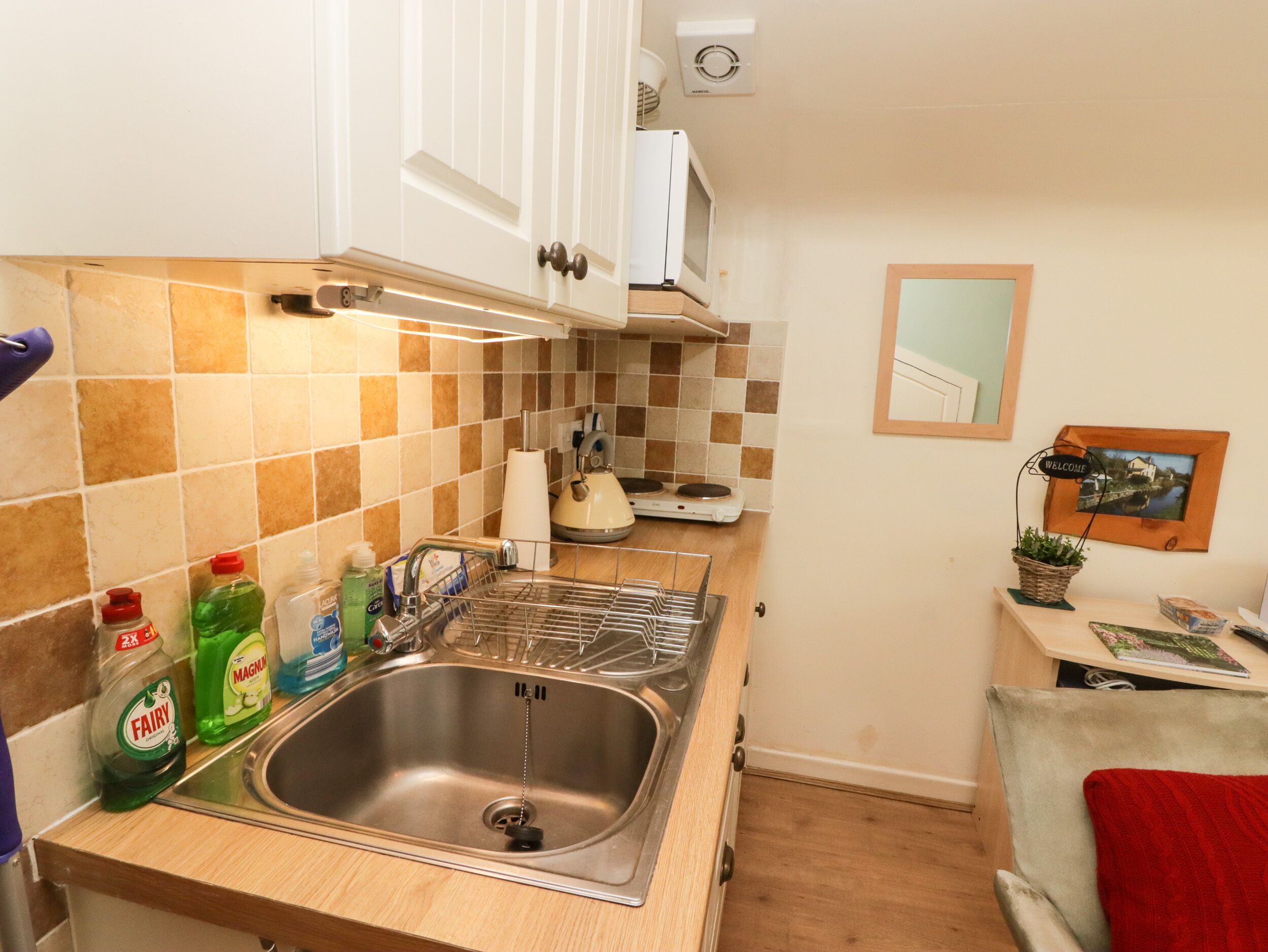 A compact kitchen with a sink, stove, and microwave. There are cleaning supplies on the counter, and a welcome sign on the wall.