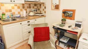 A compact kitchen with a microwave, kettle, and sink. There is a chair with a red cushion and a small table with a basket and a remote control.