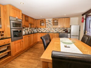 A kitchen with wooden cabinets, a dining table with chairs, and a refrigerator. - Raceside Cottage (Millom)
