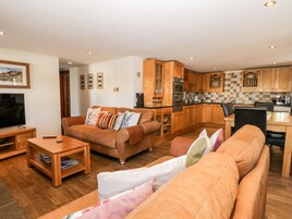 A modern living room with a brown leather sofa, a wooden coffee table, and a flat-screen TV. The kitchen features wooden cabinets and a checkered backsplash.