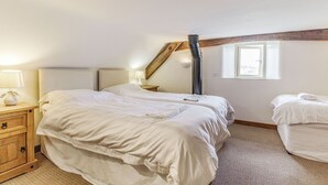 A bedroom with two beds, a wooden nightstand with a lamp, a window, and a sloped ceiling. - The Rumblings 3 (Dronfield)