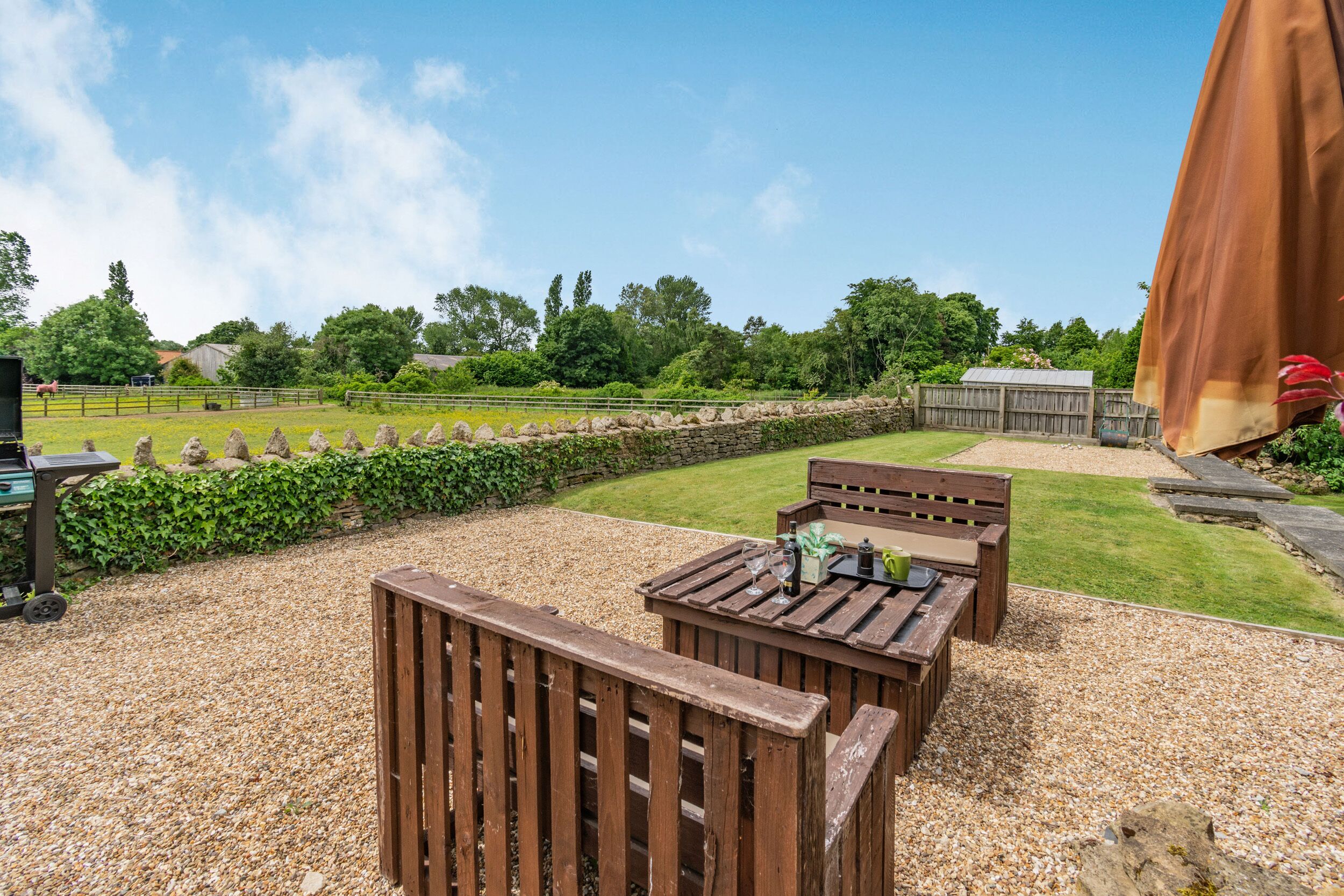 An outdoor seating area with wooden furniture and a gravel ground, surrounded by a stone wall and greenery.