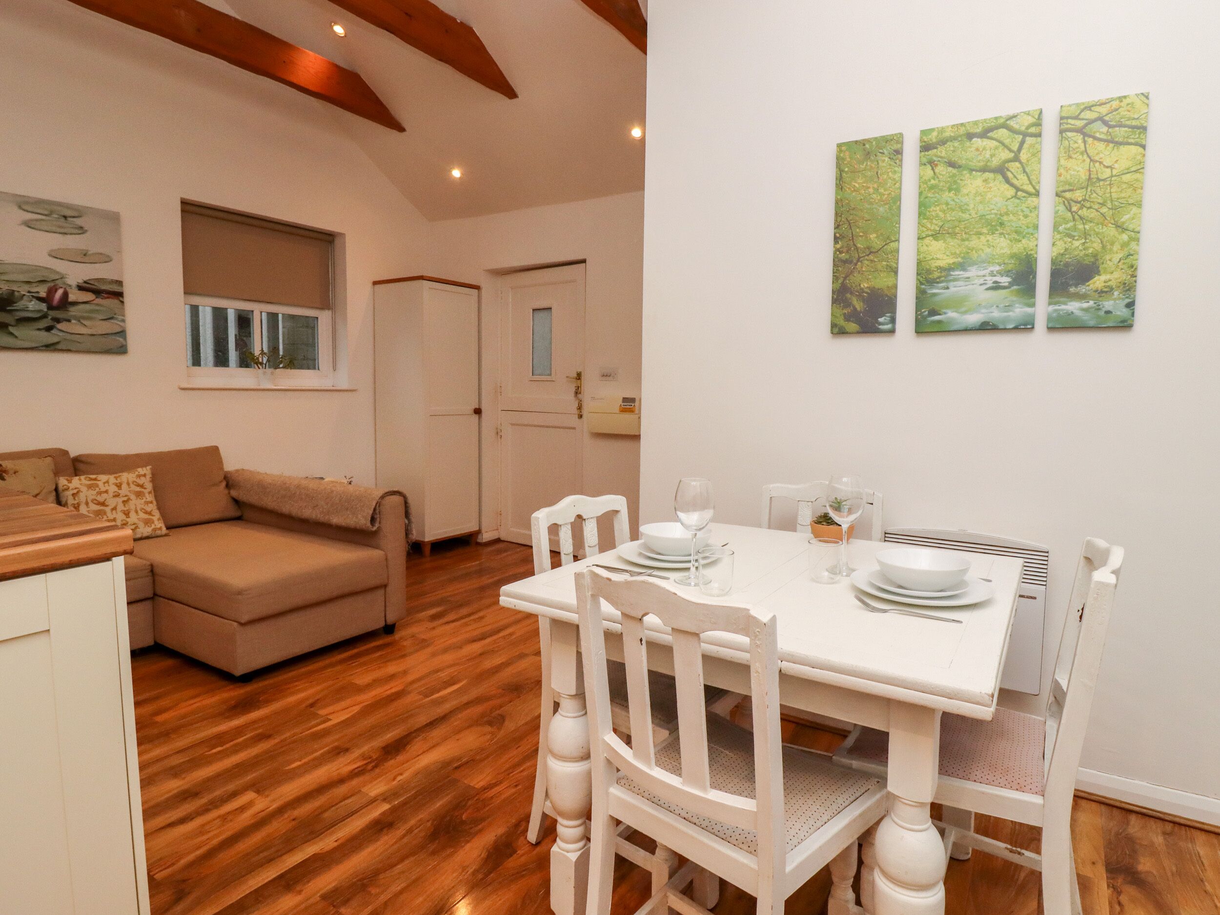 Un coin repas avec une table blanche dressée pour quatre personnes, entourée de chaises blanches. Un canapé beige est placé contre le mur, et une fenêtre avec des stores est visible.