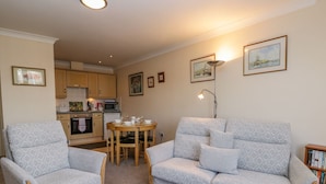 Una sala de estar con dos sillones estampados, un sofá, una mesa de centro redonda y una mesa de comedor con sillas. Se observa una cocina al fondo con armarios y electrodomésticos.