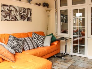 A living room with an orange sofa, patterned cushions, a floor lamp, and a glass door leading to another room. - Sunningdale (Ludlow)