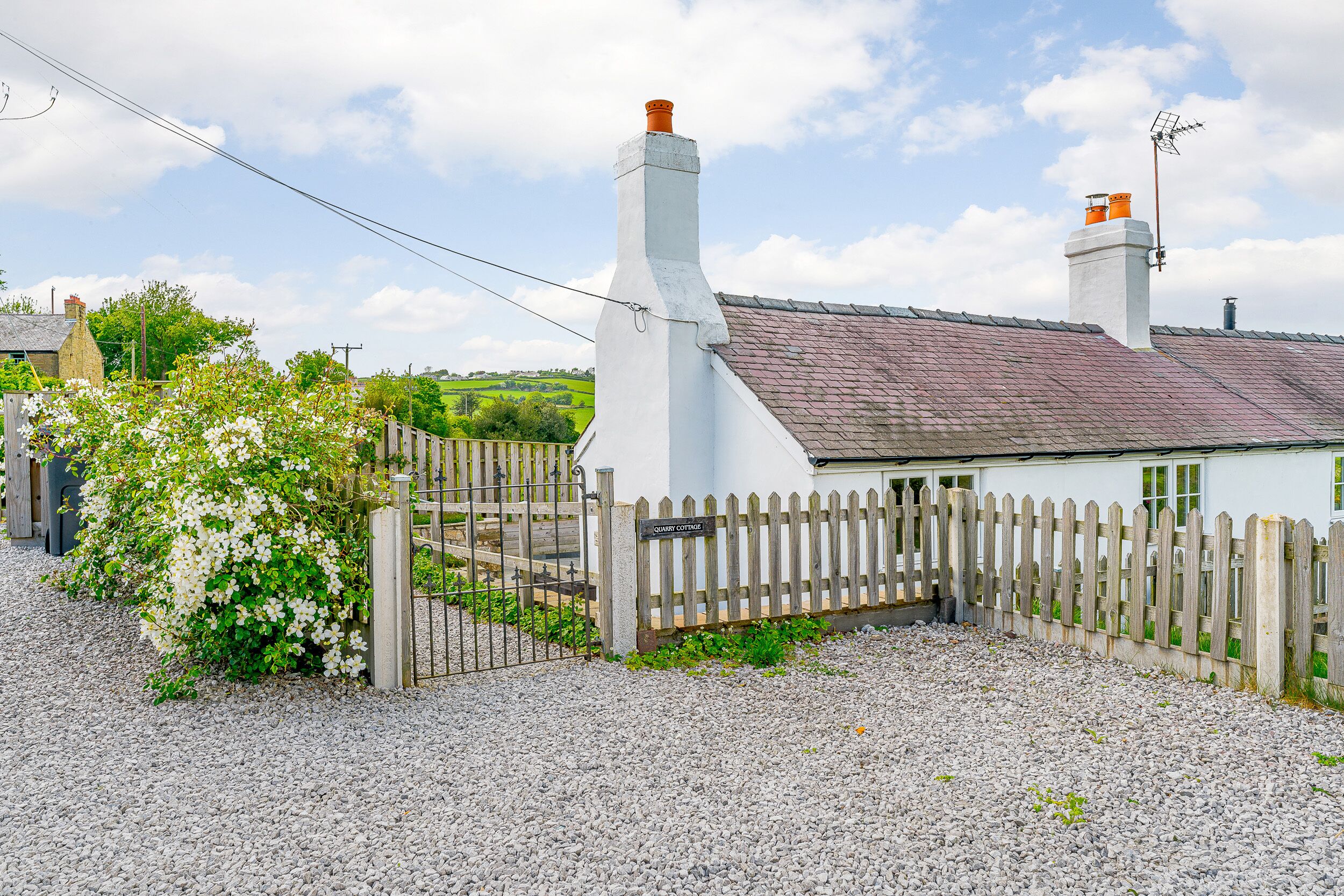 A quaint cottage with a white exterior, a wooden fence, and a gravel driveway.