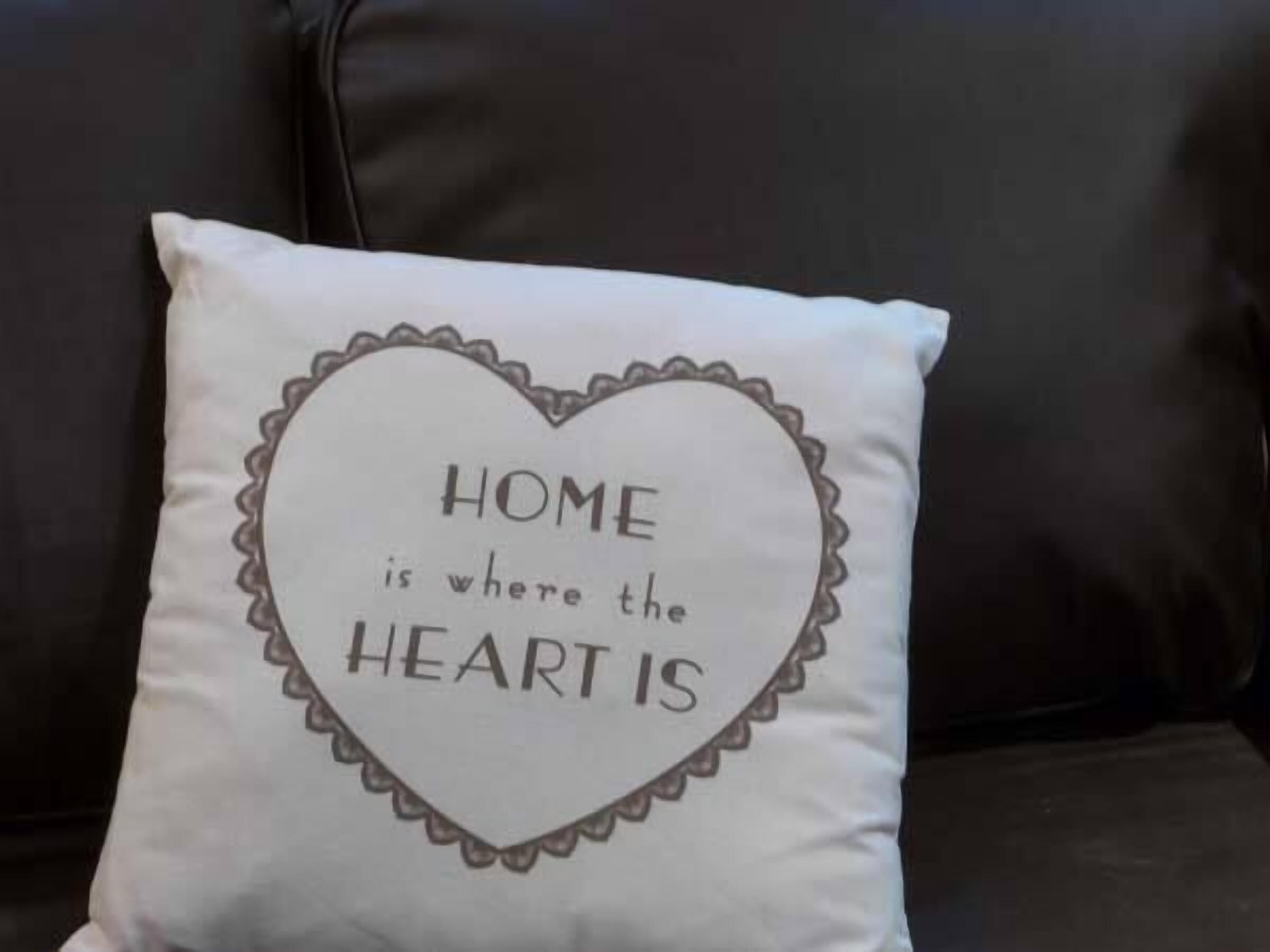 A pillow with the text "HOME is where the HEART is" on a dark-colored sofa.
