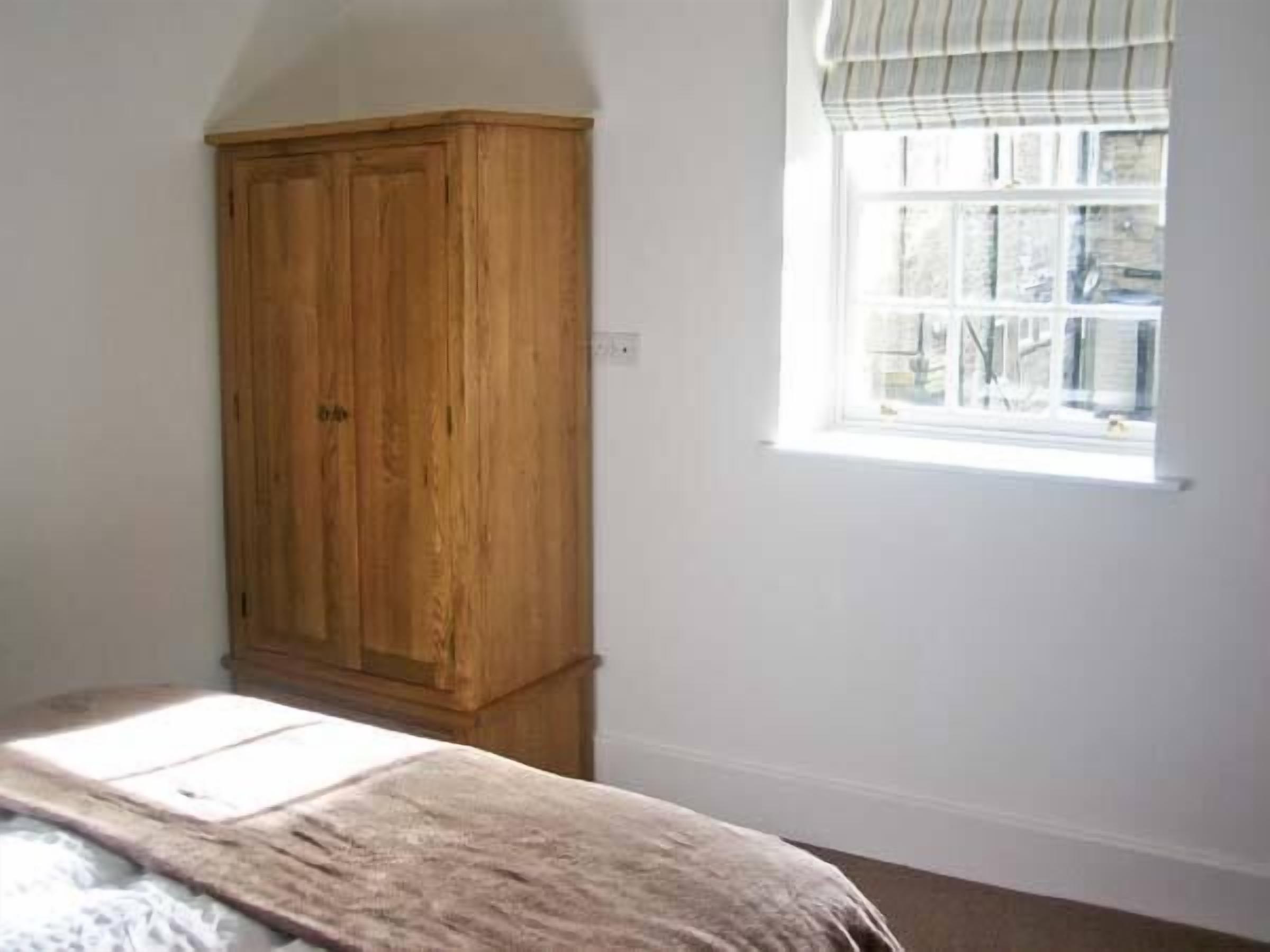 A bedroom with a wooden wardrobe, a window with blinds, and a bed with a white sheet.