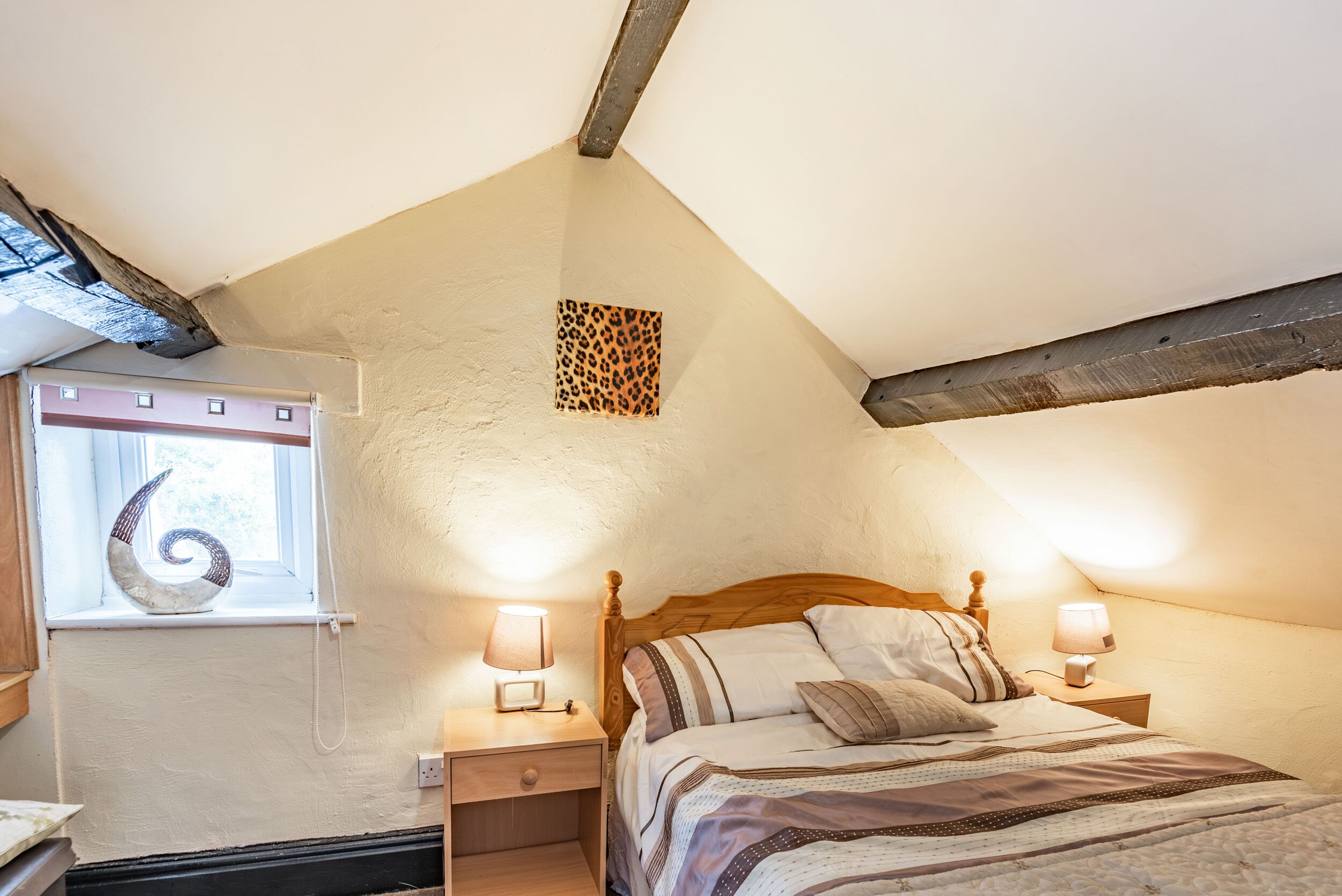 A bedroom with a sloped ceiling, a window with a decorative item, a bed with a striped bedspread, and two bedside lamps.