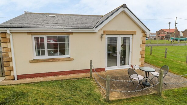 A small, single-story building with a beige exterior, a white window, and a glass door. There is a small patio with a table and chairs. - White Rock Cottage (Chathill)