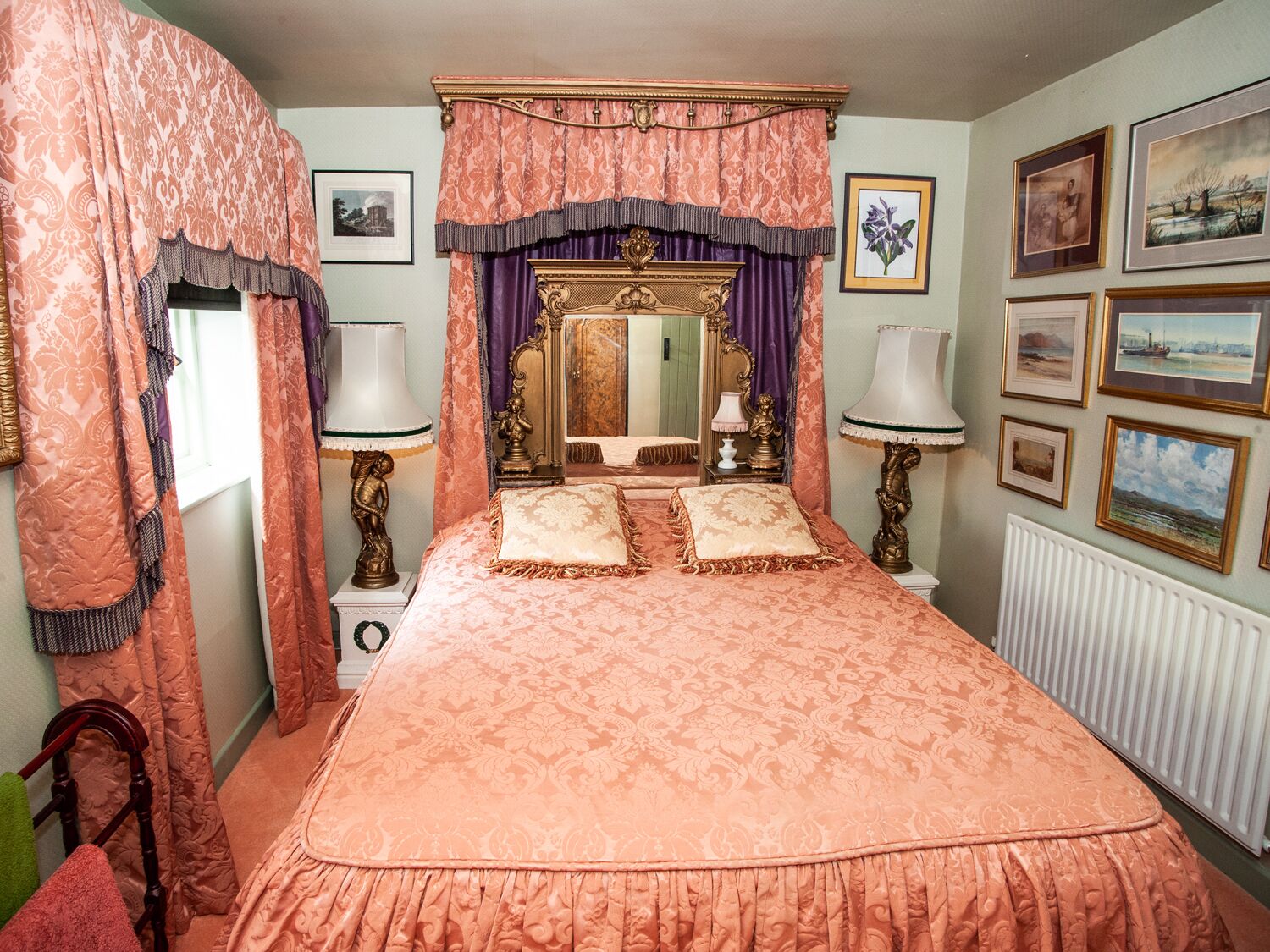 Ein Schlafzimmer mit einem großen Bett, verzierten Vorhängen, einem Spiegel und gerahmten Bildern an der Wand.