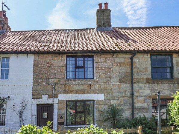 A stone house with a tiled roof, a chimney, and a small garden with plants.