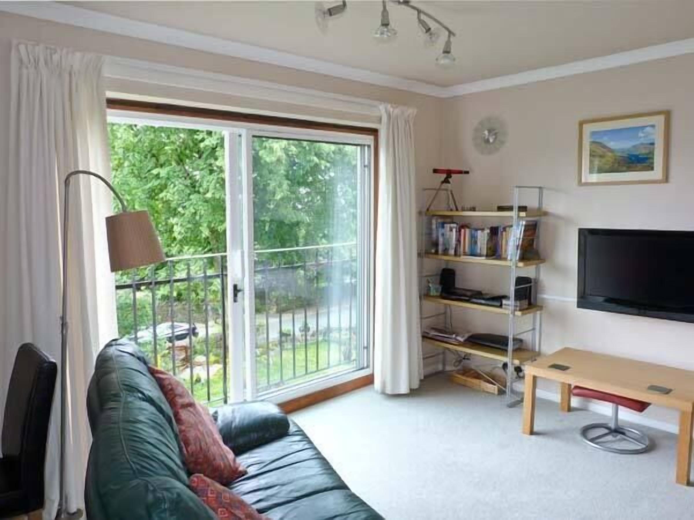 Een woonkamer met een bank, een televisie, een boekenkast en een balkon met gordijnen.