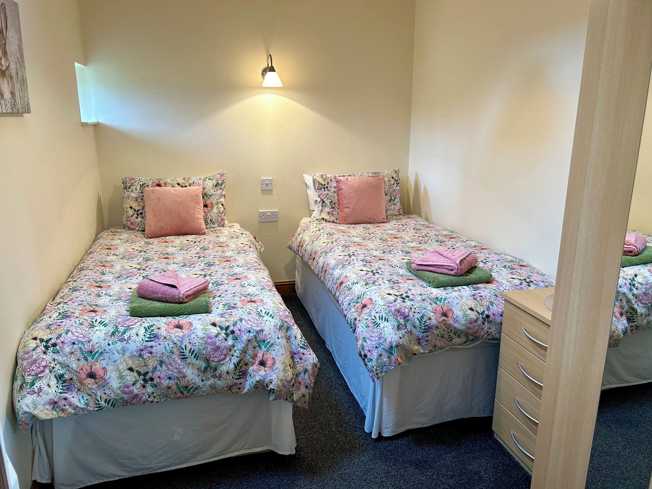 Habitación doble con dos camas, cada una con ropa de cama floral y almohadas rosas, una mesita de noche con lámpara y un aparador de madera.