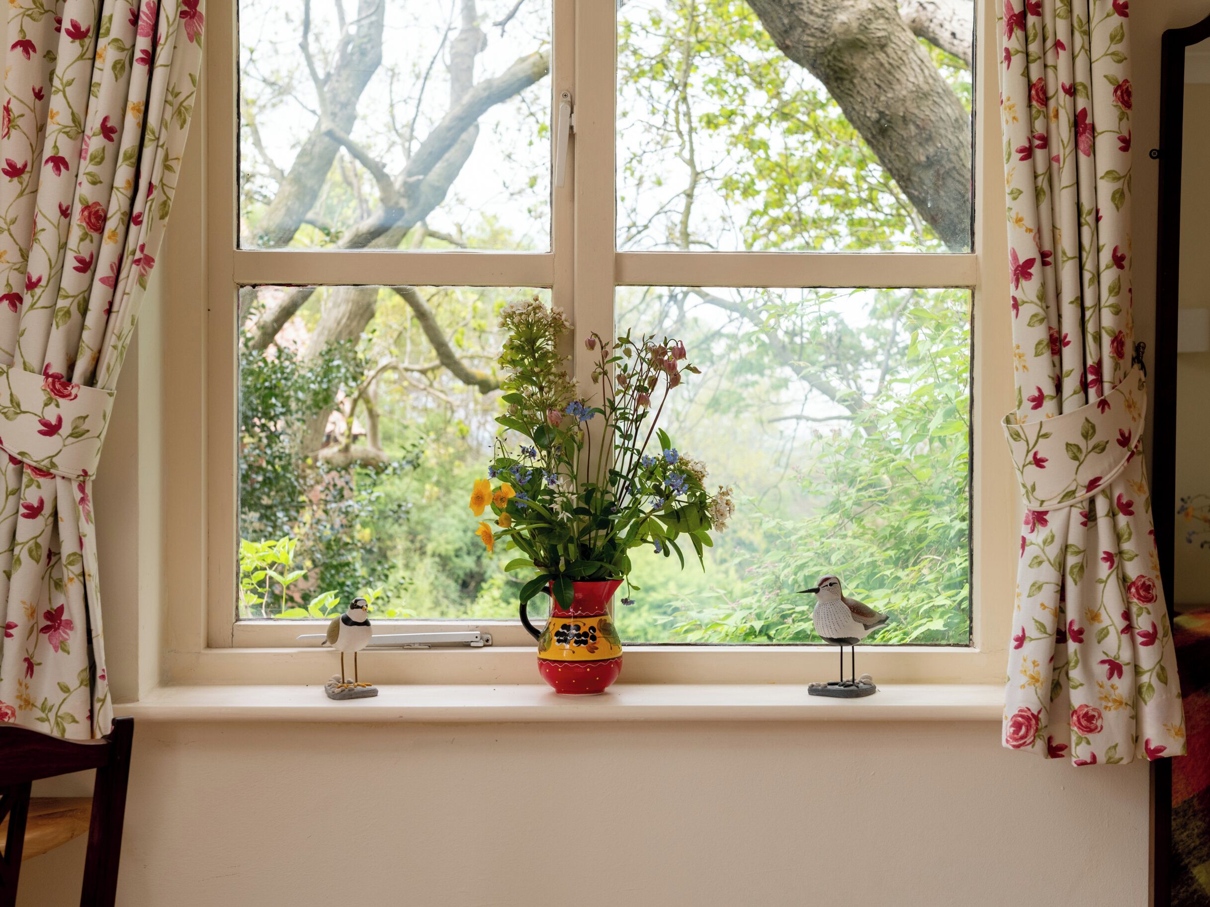A window with floral curtains, a vase with flowers, and two bird figurines on a windowsill.