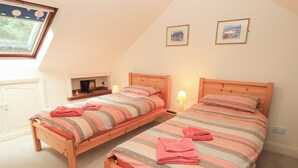 A bedroom with two wooden beds, each with a striped bedspread and pink towels. There are bedside lamps, a small table with a lamp, and a window with a view of greenery. - Uplands (Sherborne)