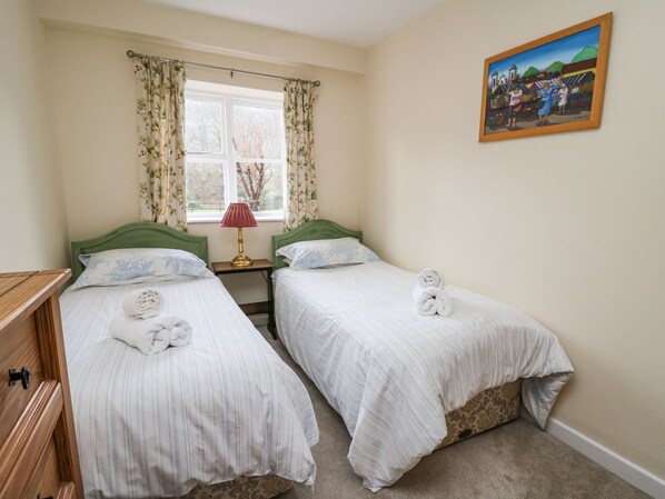 Quarto duplo com duas camas, uma janela com cortinas florais, mesa de cabeceira com abajur e um quadro na parede.