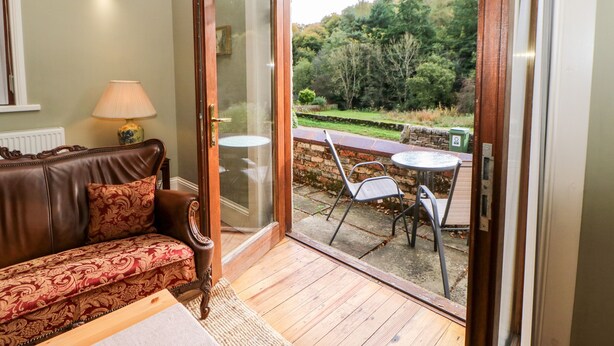 A cozy room with a brown leather sofa, a small table with a lamp, and a glass door leading to an outdoor patio with a table and chairs. - Leadmill House Workshop (Bishop Auckland)