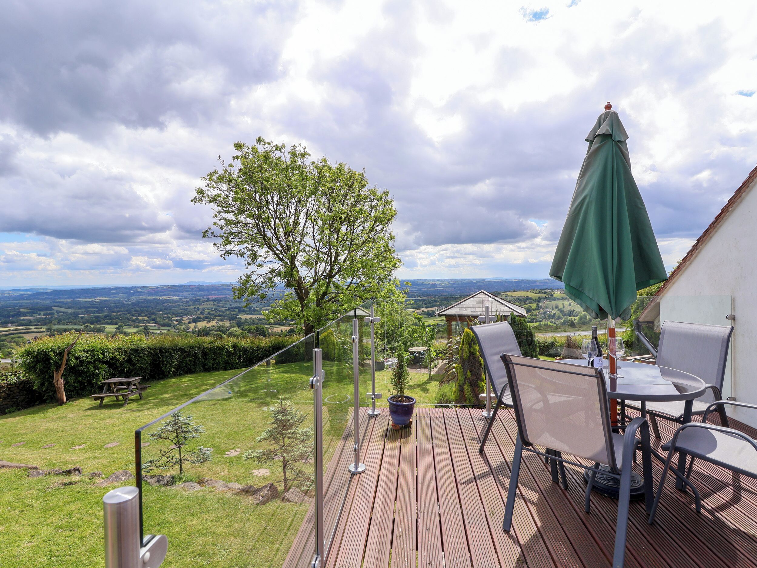 A deck with a table and chairs, overlooking a lush garden and a scenic view of rolling hills.