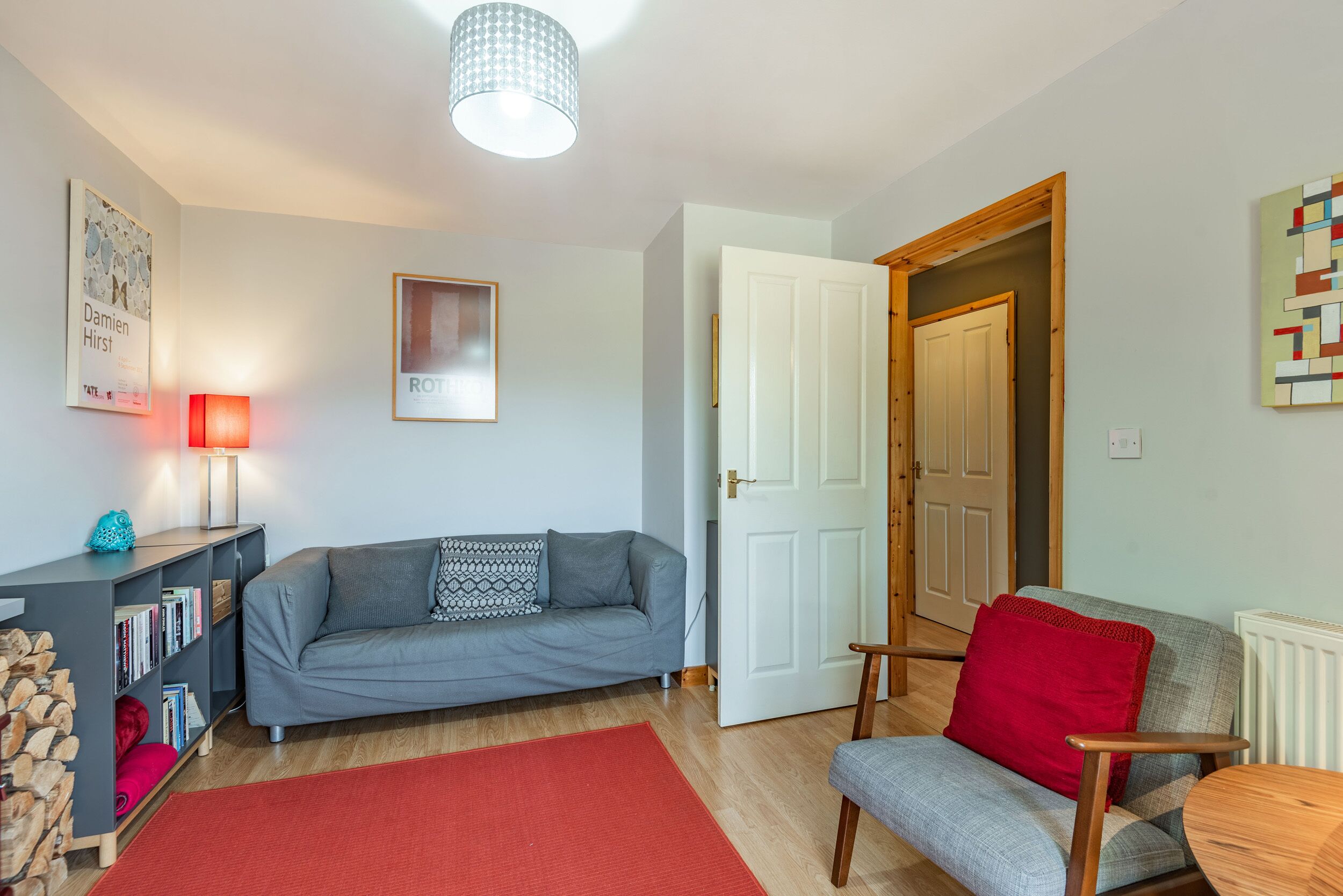A living room with a grey sofa, a red rug, a bookshelf, and a door leading to another room.