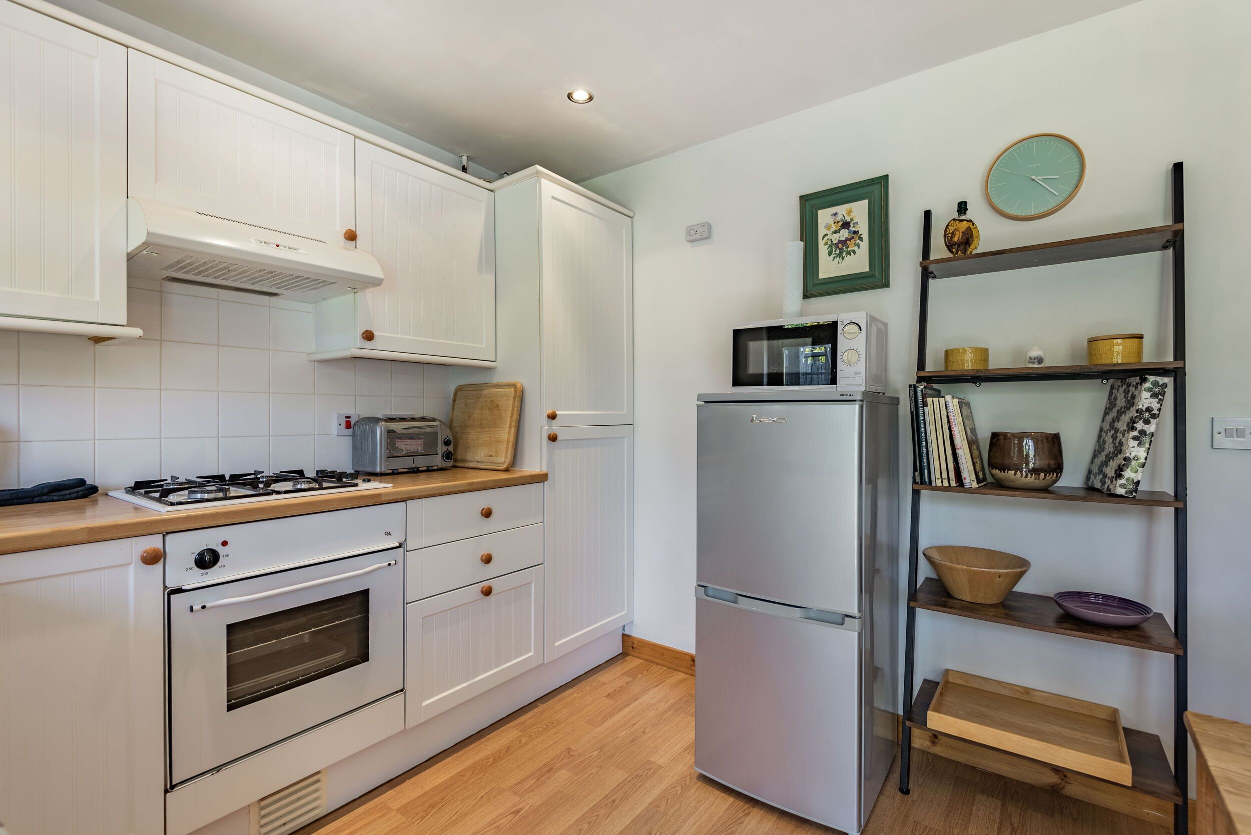 A compact kitchen with white cabinetry, a gas stove, a built-in oven, a microwave, and a refrigerator. There are wooden shelves with decorative items and a clock on the wall.