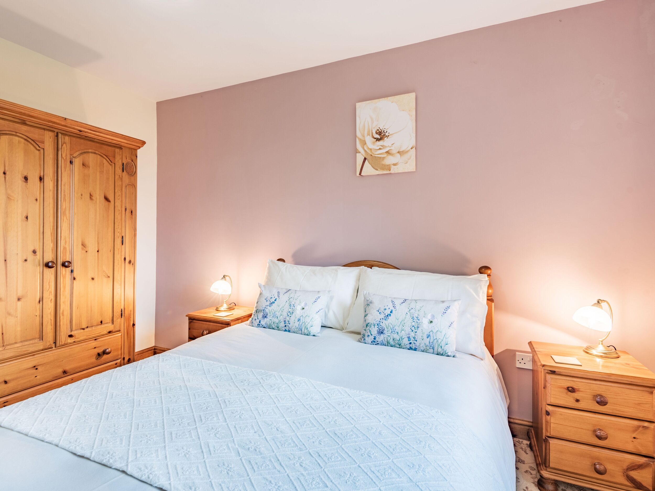 Une chambre à coucher avec un grand lit, deux tables de chevet avec des lampes, une armoire en bois et un tableau floral accroché au mur.