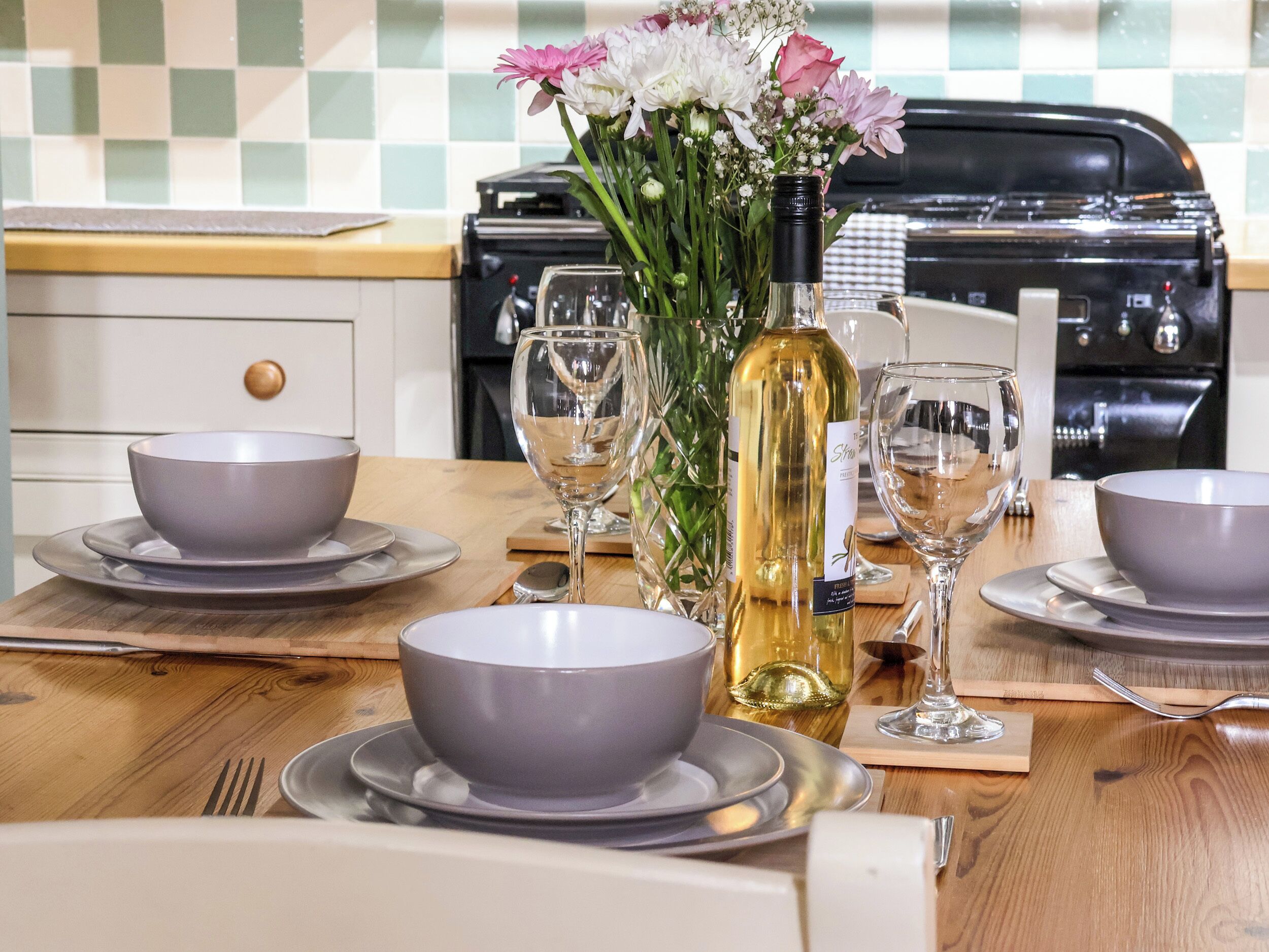 A dining table set with bowls, plates, and wine glasses, a bottle of wine, and a vase with flowers.