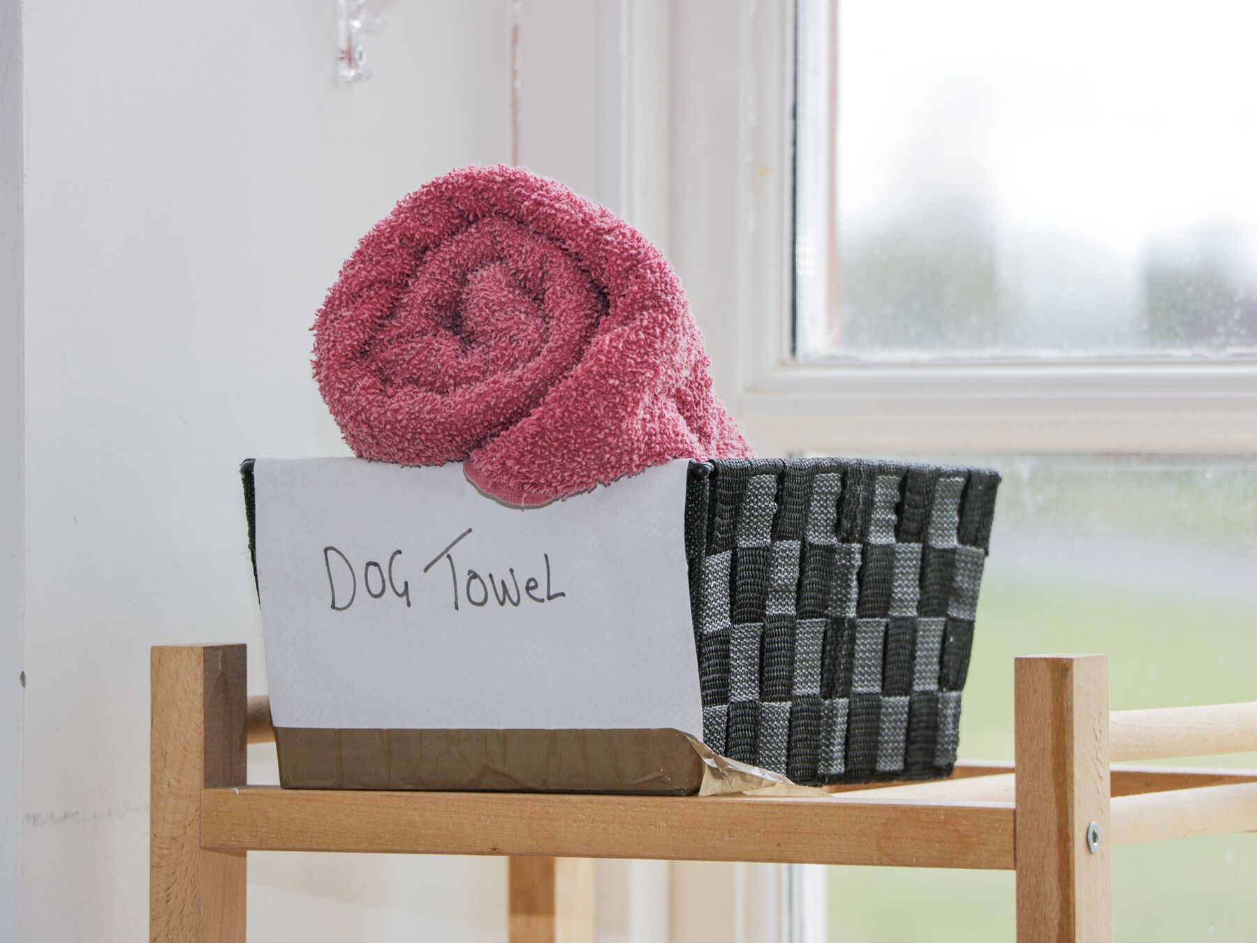 Una toalla para perro sobre una caja blanca con la etiqueta "Dog Towel" y una cesta tejida en una silla de madera.