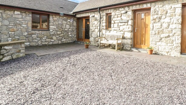 A stone building with two wooden doors and windows, a gravel courtyard, and a bench. - Cwt Blawd (Beaumaris)