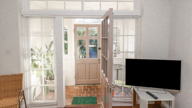 A room with a TV on a stand, a wicker chair, and a door leading to an outdoor area with a brick pathway. - Ashfield Studio (Market Rasen)