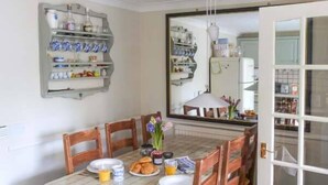 Un coin repas avec une table dressée pour quatre personnes, entourée de chaises en bois. Un miroir reflète la pièce, et une étagère contenant divers objets est fixée au mur.