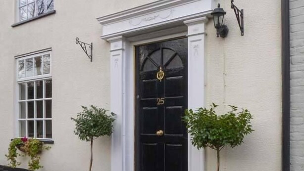 Un immeuble avec une porte noire portant le numéro 25, des moulures blanches autour de la porte et deux plantes en pot placées sur les marches.