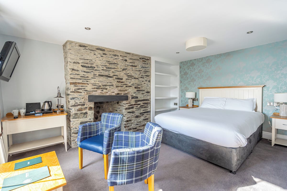 Ett hotellrum med en vägg av natursten, en säng, ett skrivbord med en bärbar dator och två fåtöljer med rutigt tyg.
